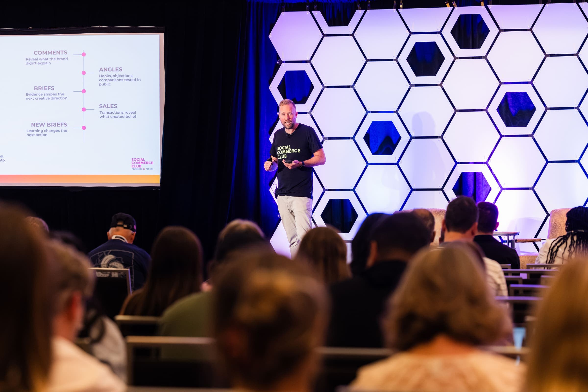 A speaker in a Social Commerce Club t-shirt presents on stage at Ecom Elevated 2026, with the audience in the foreground.